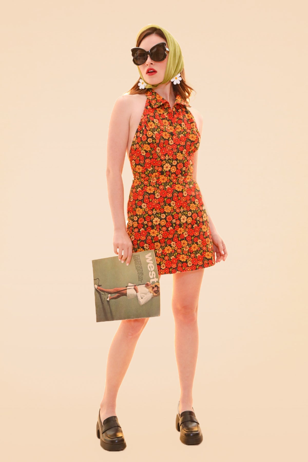 Woman wearing a colorful floral halter mini dress with sunglasses and a green headscarf holding a magazine in black loafers by Miracle Eye