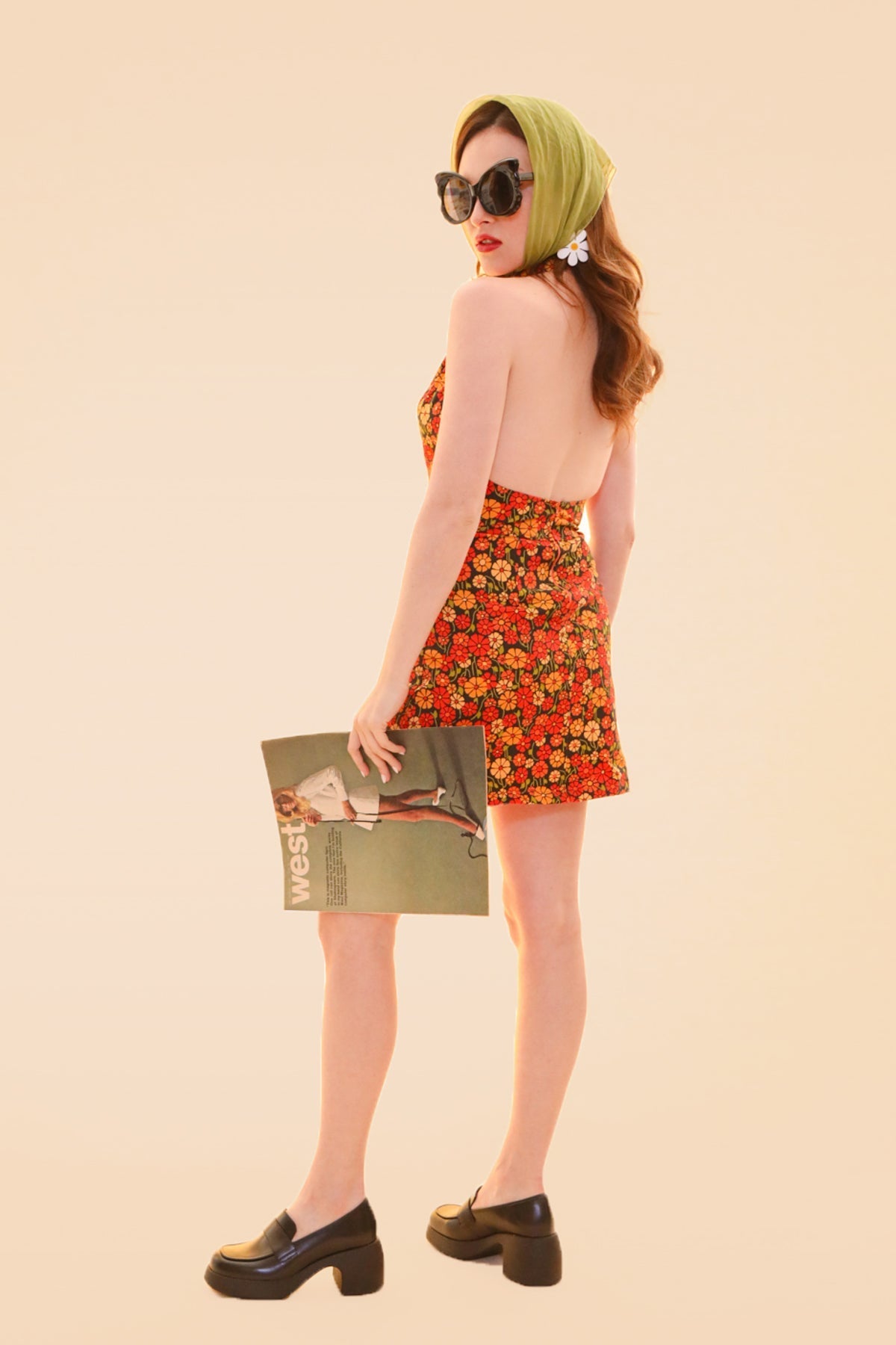 Model wearing the Miracle Eye Cecilia mini dress with a colorful floral print and open back styled with sunglasses and black platform shoes while holding a magazine