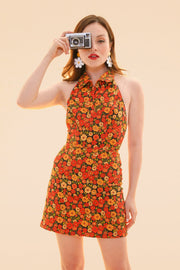 Model in a marigold floral halter mini dress with a flared skirt holding a vintage camera against a peach backdrop from Miracle Eye New Arrivals