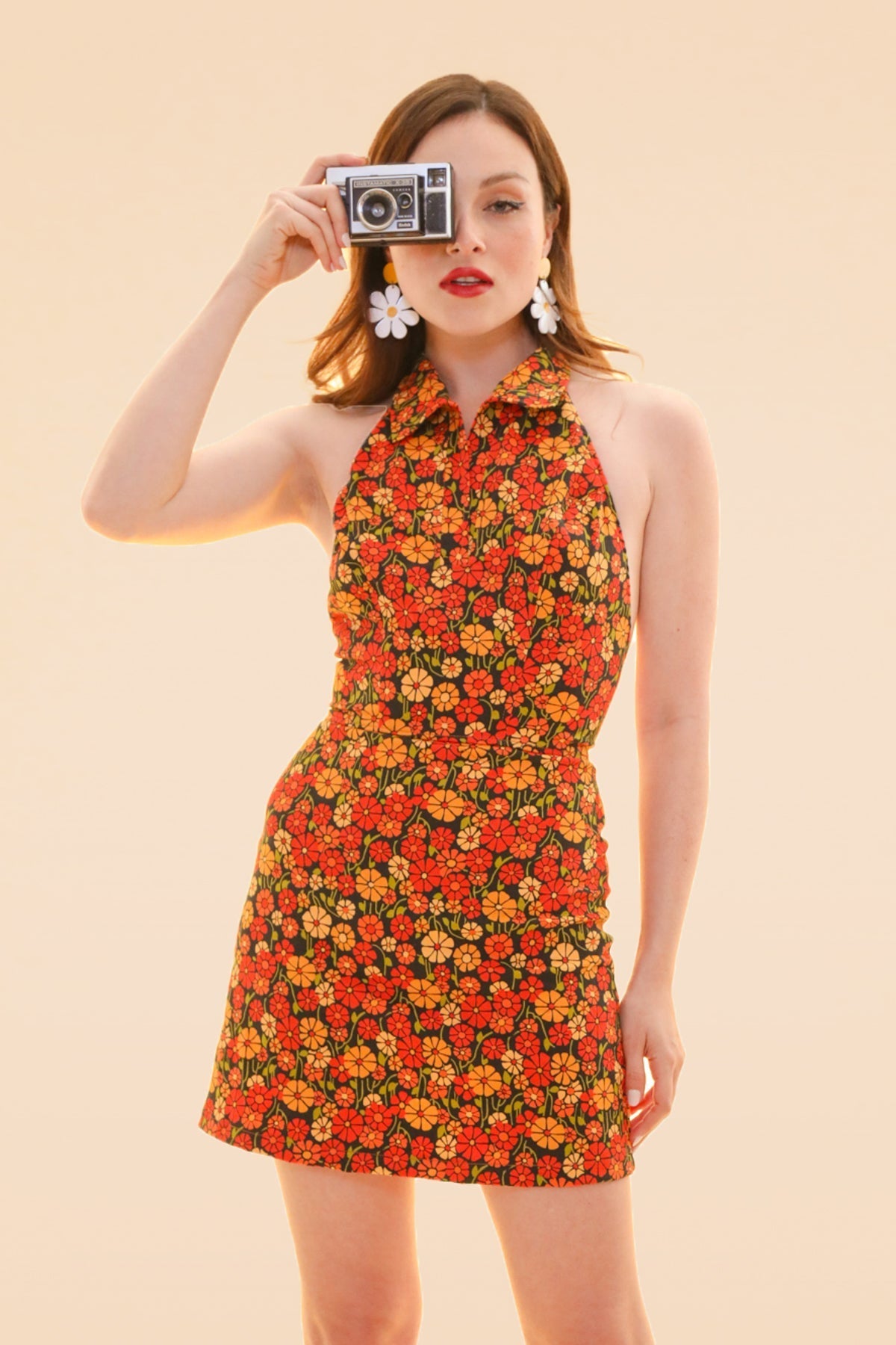 Model in a marigold floral halter mini dress with a flared skirt holding a vintage camera against a peach backdrop from Miracle Eye New Arrivals