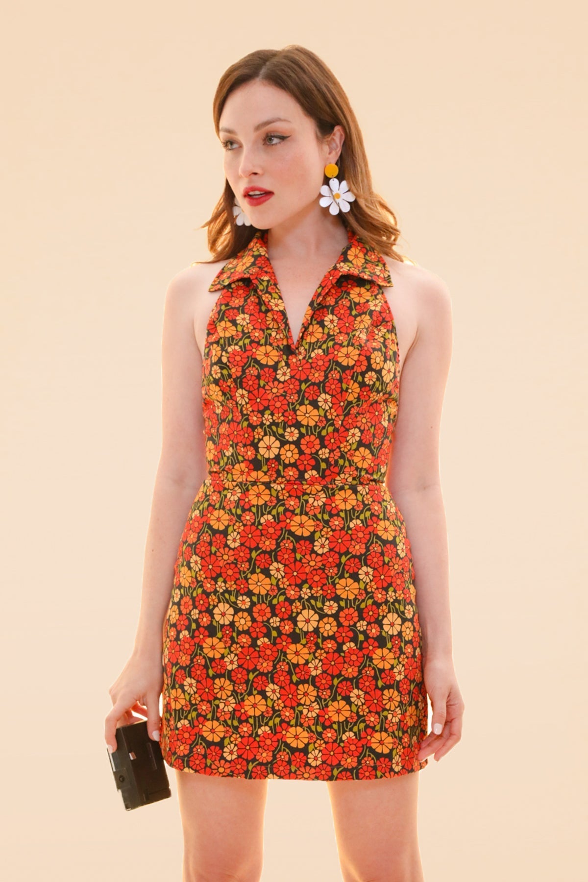 Woman wearing a Miracle Eye floral mini dress with a collared neckline and sleeveless cut while carrying a black handbag against a warm peach background