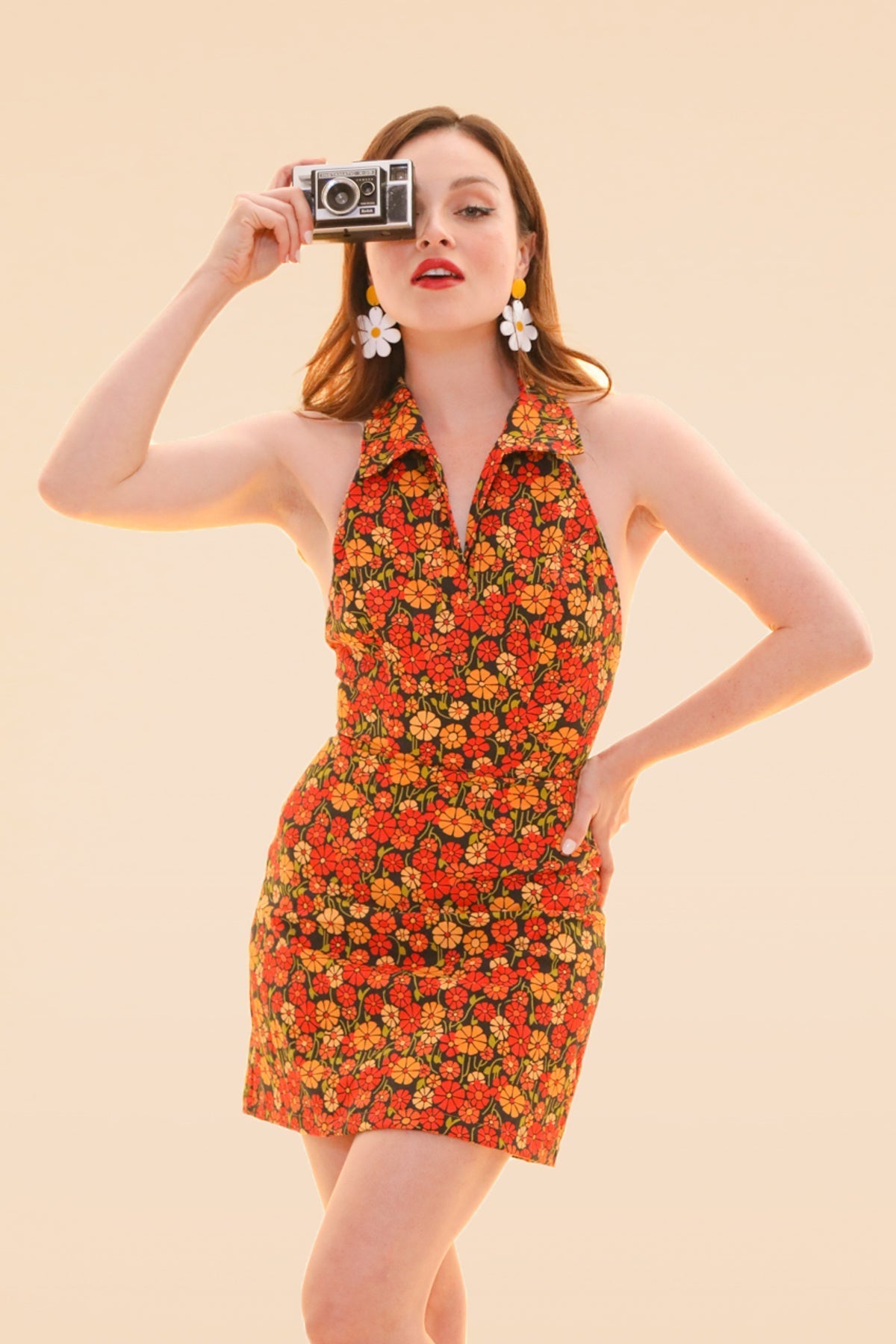 Woman in a marigold floral halter mini dress holds a vintage camera in a bright studio image from Miracle Eye New Arrivals and Sustainable Dresses and One Pieces