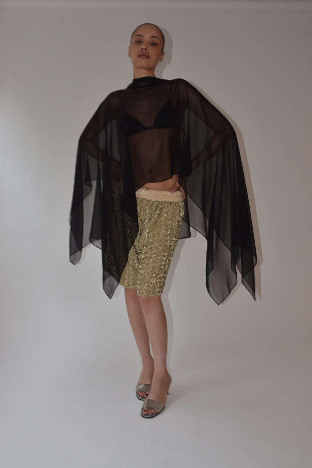Model wearing a sheer black silk chiffon caftan over a black crop top with a textured gold skirt and strappy heels by Kat Zarra in New Arrivals