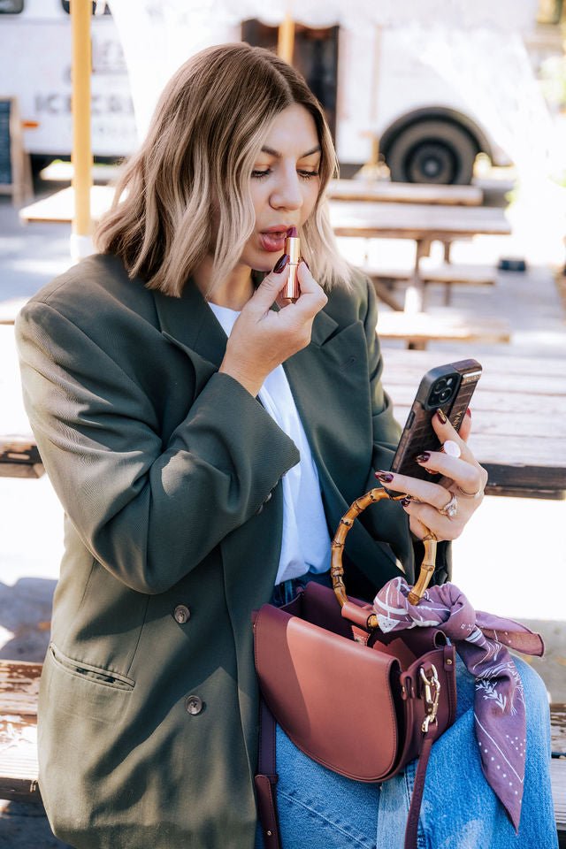 Margo Paige burgundy vegan semi circle crossbody bag styled with a patterned scarf as a woman applies lipstick outdoors