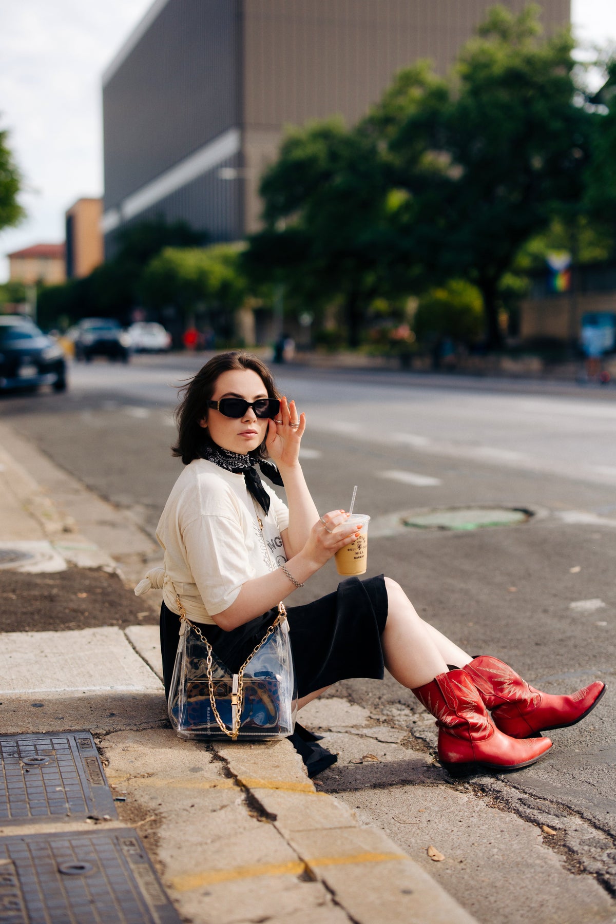 Woman seated on a city sidewalk wearing sunglasses and red boots with a clear crossbody bag and gold chain strap from Margo Paige New Arrivals collection