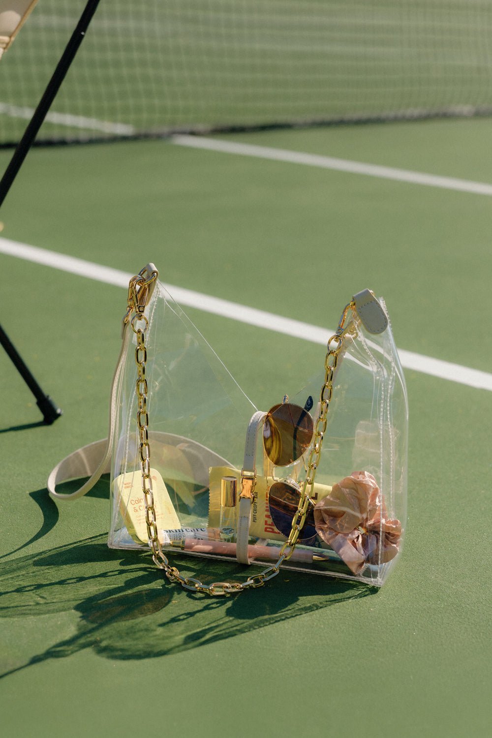 Clear cut out crossbody bag with gold chain resting on a tennis court with visible essentials inside from Margo Paige New Arrivals collection